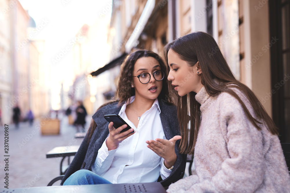 Φωτογραφία two beautiful stylish women sitting at the table in street cafe, looking at the phone and talking
