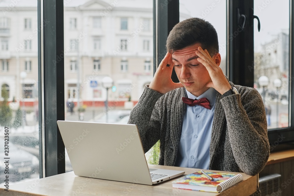 Fototapeta premium Young tired, ill, overworked man in formal wear sitting in front of computer and touching his forehead in the cafe