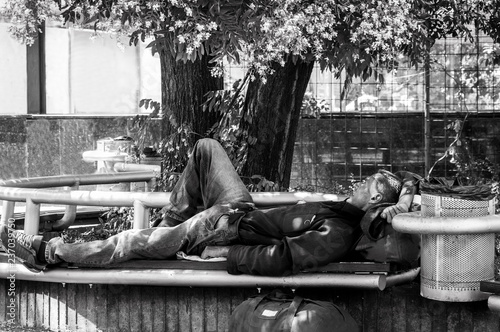 Poor hungry and tired homeless veteran man ex military soldier sleep in the shade on the bench in urban city street social documentary concept black and white hard contrast