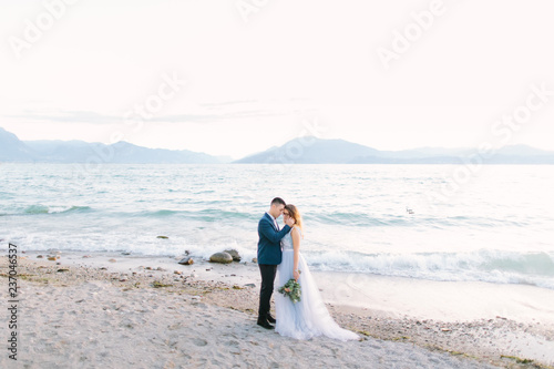 Tender portrait of young man hugging his pretty woman while standing on the pier near the lake on the wedding ceremony. Garda Lake  Italy