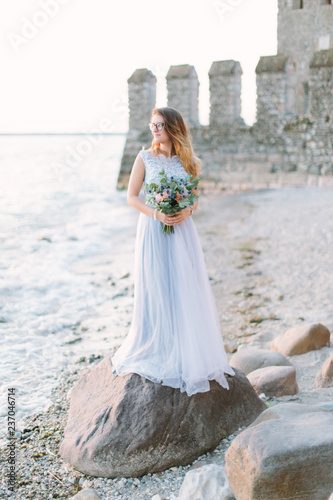 A charming young girl in a wedding blue dress standing on rocks near Rocca Scaligera Castle in Sirmione. Garda Lake