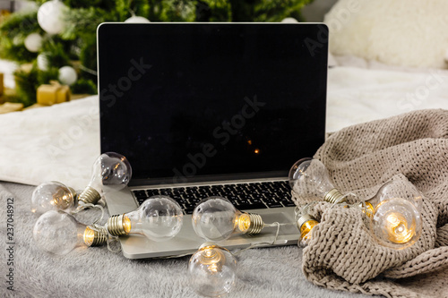 Image of open laptop with white screen on wooden table in front of christmas tree background. For mockup