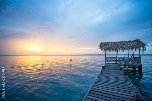 A bird is flying in the scene showing a wooden dock with a palapa roof and sunset. The water is calm and blue. photo
