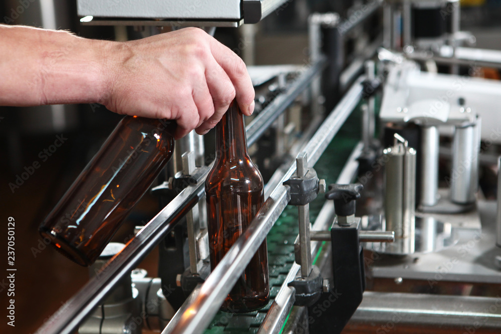 Hand worker puts an empty beer bottle on the conveyor of the plant for the production of beer. Only the bottle is in focus. Man's hand close-up.The concept of production. Hand worker puts an empty beer bottle on the conveyor of the plant for the production of beer. Only the bottle is in focus. Man's hand close-up.The concept of production.