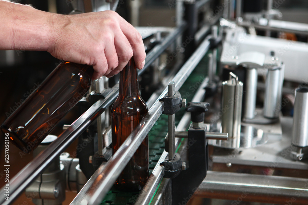 Hand worker puts an empty beer bottle on the conveyor of the plant for the production of beer. Only the bottle is in focus. Man's hand close-up.The concept of production.