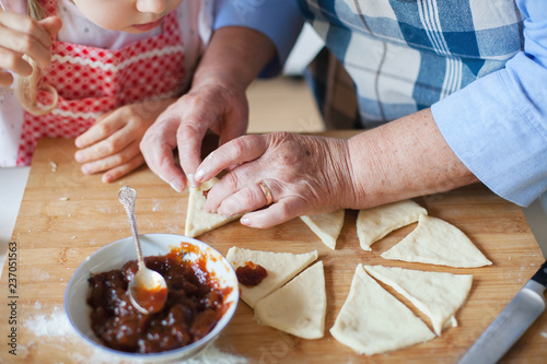 Grandmother is teaching granddaughter to cook croissants. Hands of child and old retired woman are baking in home kitchen. Senior woman and kid girl are preparing for family Thanksgiving Day dinner photo