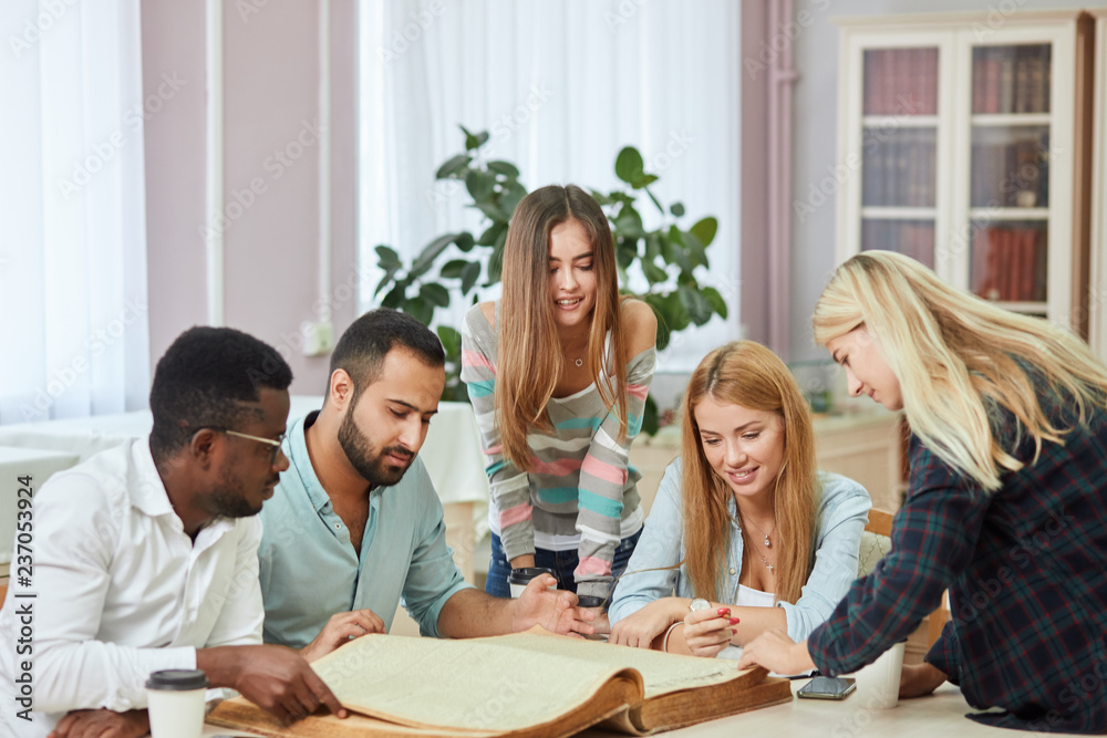 Pensive african american young man in stylish eyeglasses for vision correction, Arab man and three european young female students reading big old textbook sitting at the table against window .
