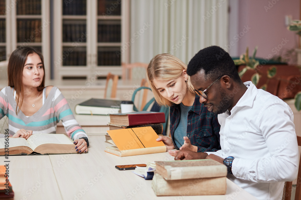 Diverse curious adult students learning together, reading old large reference book in library, deepen their knowledge and understanding in philology.