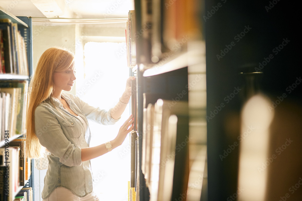 Blonde caucasian young woman searching book in the old bookstore walking along long rows of bookshelves picking out something unusual for gift to her boyfriend