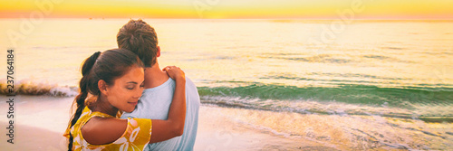 Couple in love hug banner panorama. Asian woman hugging embracing boyfriend relaxing on beach watching sunset - love and tenderness on travel summer holidays.