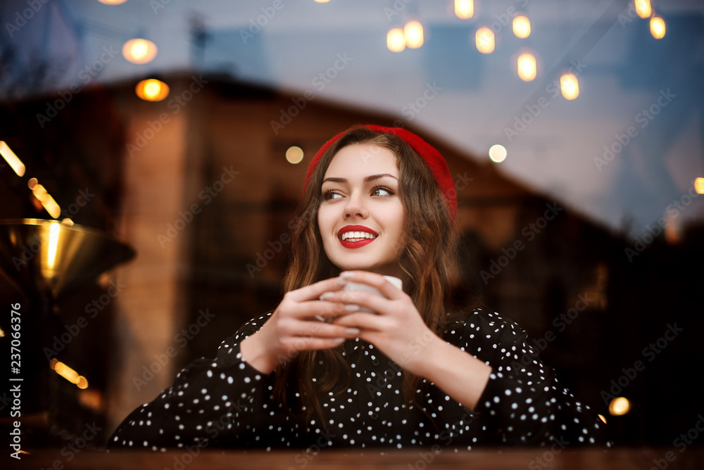 Young beautiful fashionable happy smiling woman with red lips, wearing  french style beret and polka dot blouse drinks coffee or tea in cafe with  loft interior. View through the window glass Photos