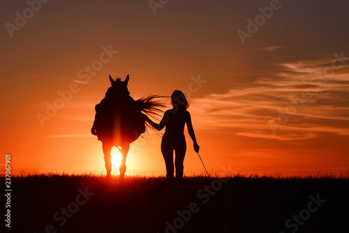 Woman and horse walk in field with red rising sun on horizon. Beautiful Idyllic and romantic sunset background with equine and girls silhouette on horse hiking