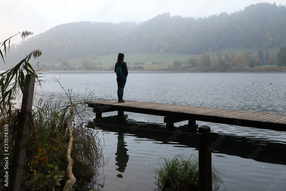 A girl standing on a pier and her reflection on a lake.