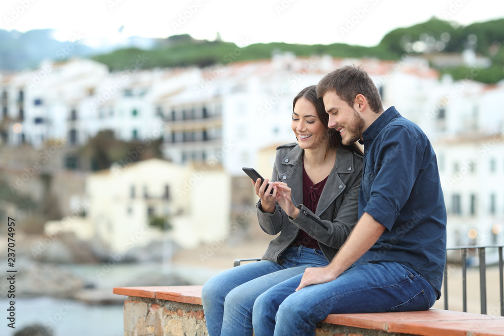 Happy couple on a ledge checking smart phone Happy couple on a ledge checking smart phone