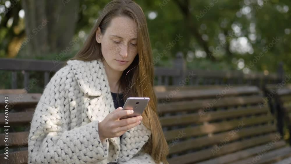 Slow motion portrait of beautiful young woman with freckles, trendy coat using smartphone, checking social networks, enjoying warm evening sitting in city park . Slow motion portrait of beautiful young woman with freckles, trendy coat using smartphone, checking social networks, enjoying warm evening sitting in city park .