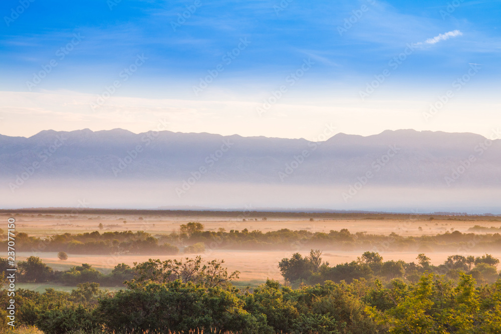 Croatia, Dalmatia, Velebit mountain ridge and misty fields in foreground, summer morning Croatia, Dalmatia, Velebit mountain ridge and misty fields in foreground, summer morning