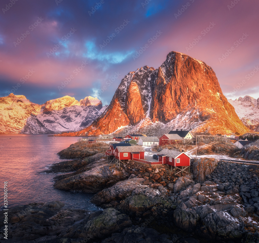 Hamnoy village on the hill at sunrise. Lofoten islands, Norway. Winter landscape with houses, snowy mountains, sea, colorful sky with clouds. Norwegian traditional red rorbu and snow covered rocks - obrazy, fototapety, plakaty 