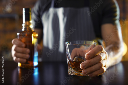 Bartender with glass and bottle of whiskey at counter in bar, closeup. Space for text