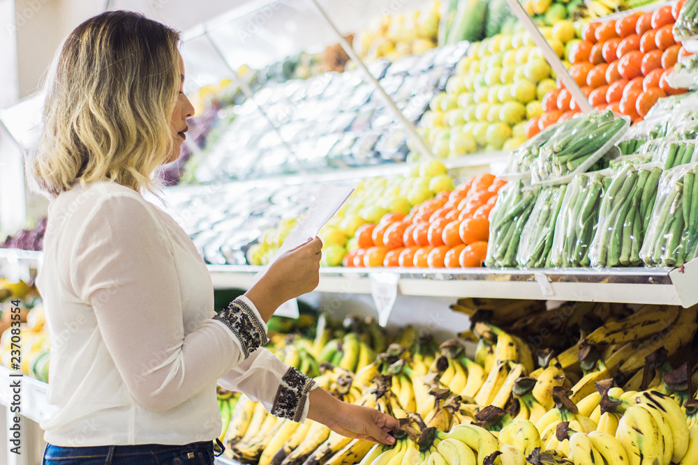 Obraz premium young woman holding shopping list with fruits background in supermarket