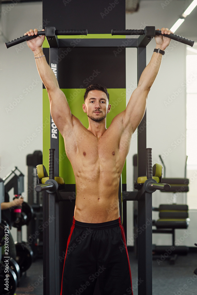 Fototapeta premium Strong attractive man does pull-up on the crossbar during training in the modern gym. Front view.