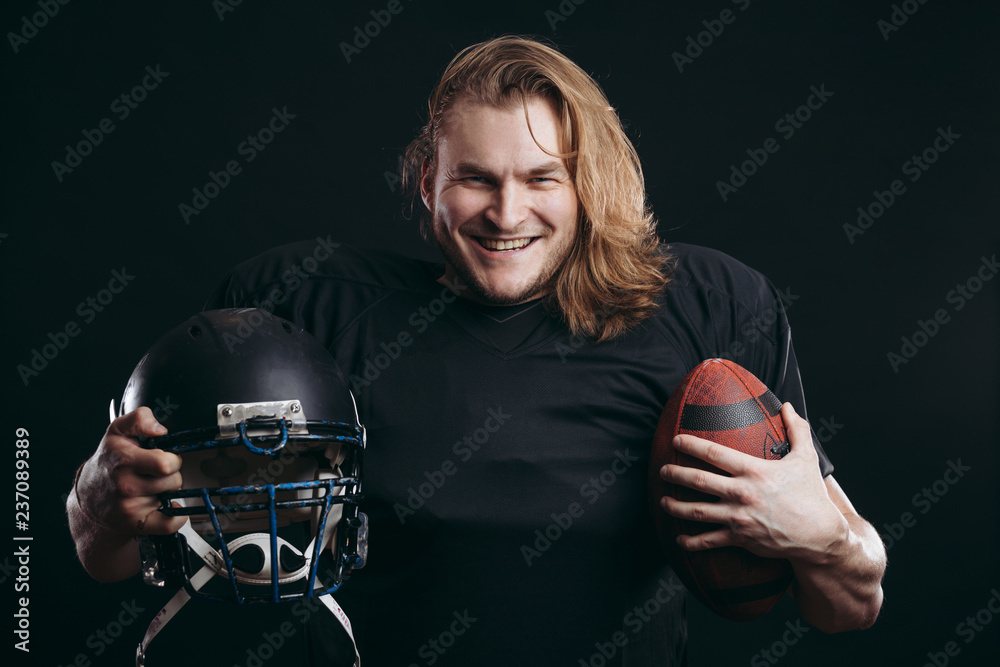 Happy american football player in black sportswear with helmet and ball over black background. People, sport, positive emotions and facial expressions concept
