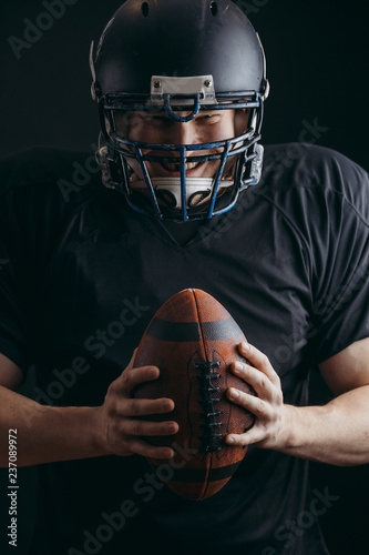 Dramatic portrait of determined American football player equipped with black headgear and protection suit, wearing oval ball isolated on black background