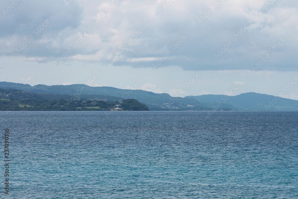 Looking across the water at the green, tree cover, hills beyond, Montego Bay, Jamaica. Water has a slight ripple. Sky is overcast.