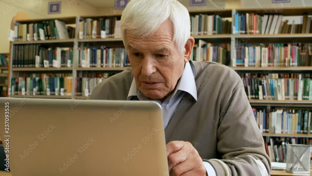 Tilt up medium shot of concentrated retired man sitting at desk in library and using laptop computer Tilt up medium shot of concentrated retired man sitting at desk in library and using laptop computer