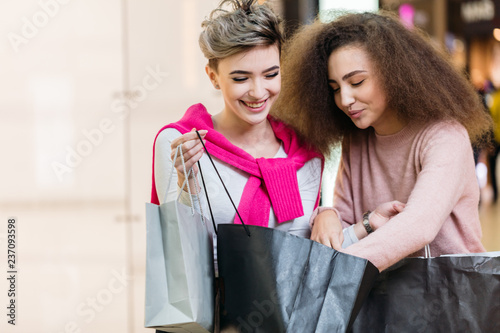 Youth, fashion, style and happiness concept. Charming young brunette female with frizzy hair showing to her blond female friend her shoppings from clothing boutique, feeling happy and excited