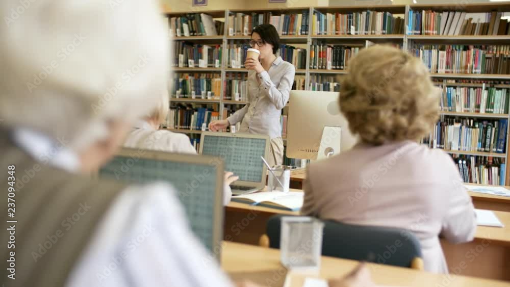 Handheld shot of young female teacher in glasses talking to group of senior people and drinking coffee during information technology lesson Handheld shot of young female teacher in glasses talking to group of senior people and drinking coffee during information technology lesson