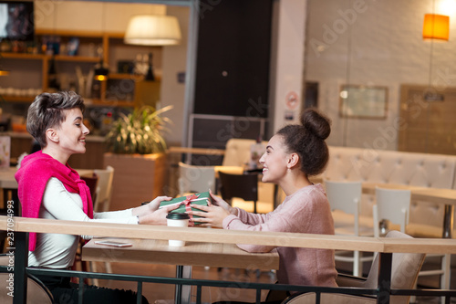 Brunette woman presents gift box to her blonde girlfriend while sitting in mall cafe. The blond girl is looking happy and surprised. Shopping, People and Gifts concept