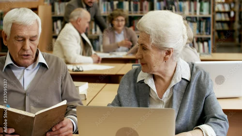 Medium shot of senior woman with laptop computer sitting at desk and talking to retired man who is reading book Medium shot of senior woman with laptop computer sitting at desk and talking to retired man who is reading book