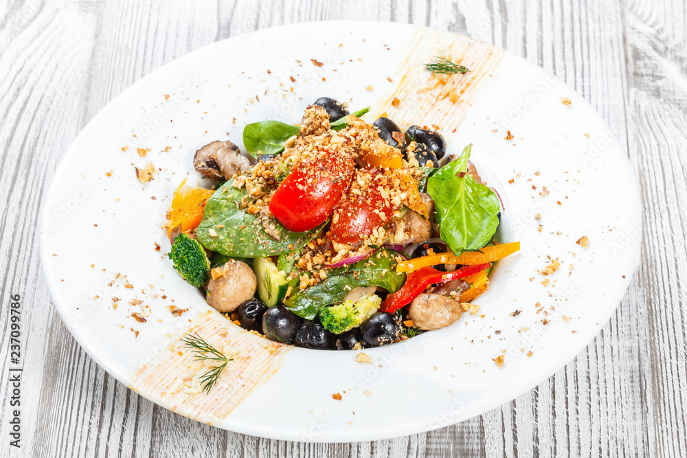 Vegetables salad with spinach, mushrooms, tomatoes, onions, cucumbers, broccoli, olives and nuts on plate on light wooden background. Healthy food. Top view