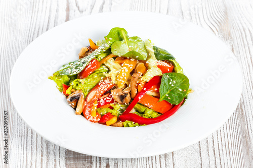 Fresh vegetable salad with lettuce, spinach, grilled mushrooms, tomatoes, sweet peppers and sesame seeds on plate on light wooden background close up. Healthy food. Top view