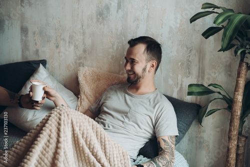 Happy man with tattooed arm lying in bed and drinking morning coffee in loft designed room © alfa27
