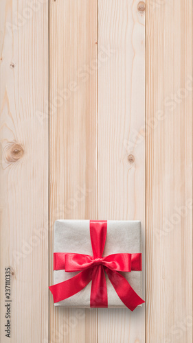 Gift box present (isolated with clipping path) with red bow satin ribbon over brown wrapping paper on white pine wood background for Christmas and birthday greeting card design decoration