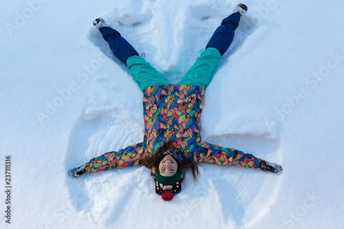 High angle view of happy girl lying on snow and moving her arms and legs up and down creating a snow angel figure. Smiling woman lying on snow in winter holiday with copy space