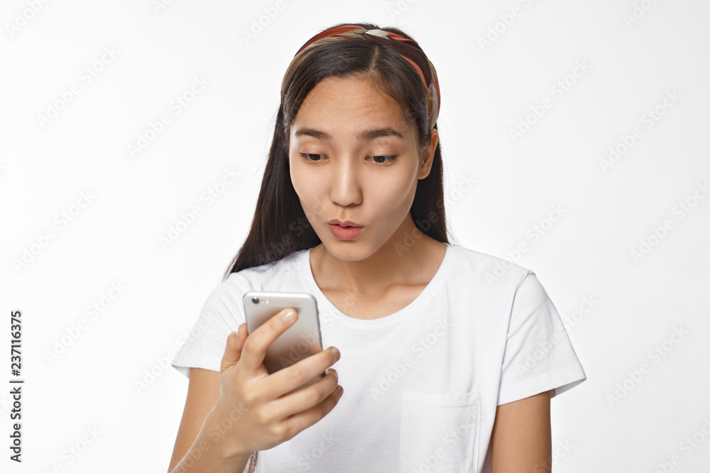Beautiful young Chinese female with black loose hair having surprised astonished look, staring at screen of generic cell phone, watching cute video, picture or reading text message, surfing internet Beautiful young Chinese female with black loose hair having surprised astonished look, staring at screen of generic cell phone, watching cute video, picture or reading text message, surfing internet