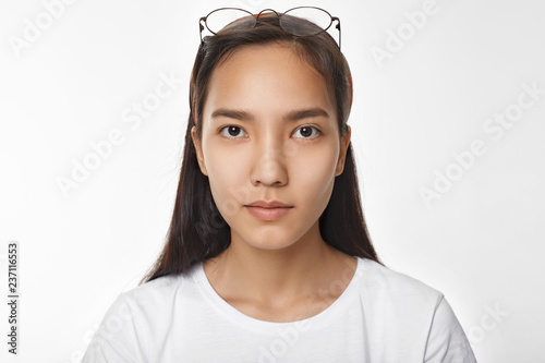 Ethnicity and races concept. Beautiful adorable teenage girl with Asian oriental eyes and tanned skin posing isolated in studio. Pretty Chinese woman wearing white top and glasses on her head