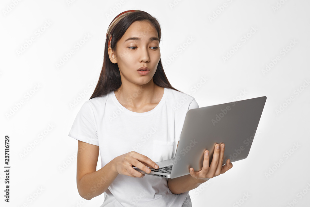Surprised amazed young Asian woman in casual t-skirt holding generic portable computer and looking at screen with astonished facial expressions, shocked with content, news or picture on internet