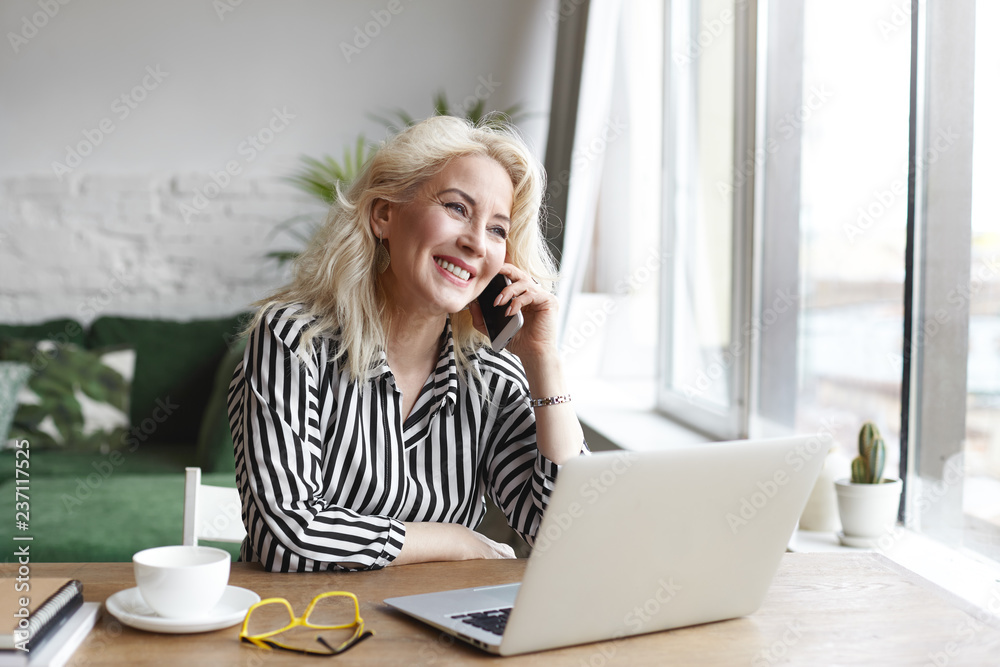 Elegant sixty year old senior self employed woman dressed elegantly, talking to client and smiling, discussing details of online purchase, sitting at wooden desk in front of open portable computer Elegant sixty year old senior self employed woman dressed elegantly, talking to client and smiling, discussing details of online purchase, sitting at wooden desk in front of open portable computer