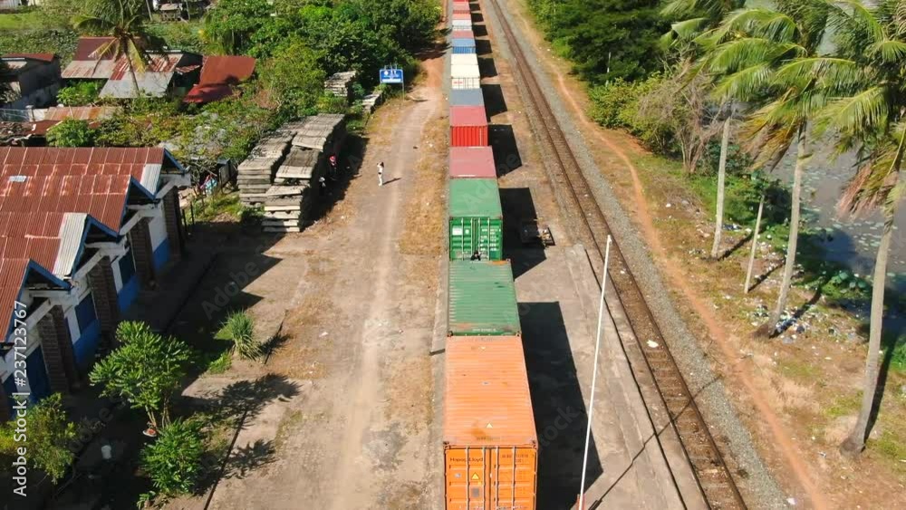 Aerial drone view fly back quickly over a parked freight train in rural tropical Southeast Asia. Camera ascends at end of shot, revealing palm trees, green ponds, and jungle background. Aerial drone view fly back quickly over a parked freight train in rural tropical Southeast Asia. Camera ascends at end of shot, revealing palm trees, green ponds, and jungle background.