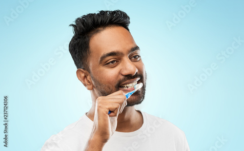 oral care, dental hygiene and people concept - smiling young indian man with toothbrush cleaning teeth over blue background