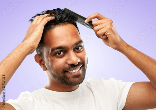 grooming, hairstyling and people concept - smiling young indian man brushing hair with comb over ultra violet background