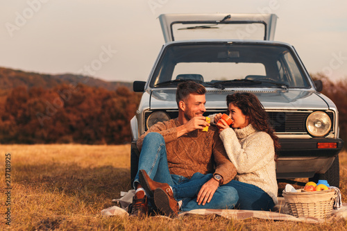 	 Beautiful young couple enjoying picnic time on the sunset. They drinking tea and sitting in a meadow leaning against a old fashioned car.