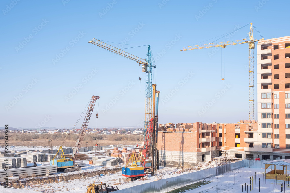 Crawler cranes, piles, concrete slabs and cranes on the construction site near the multi-storey building. The process of driving piles. Translation: 