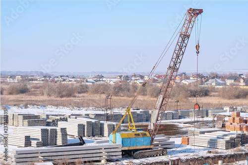 Lifting equipment. Crawler crane, piles and concrete slabs at a construction site near a multi-storey building. Translation: 