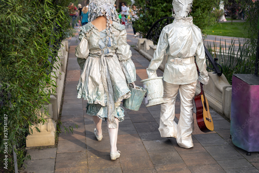 Pair of street artist painted in white silver paint walking in city park. Living statue performer with guitar