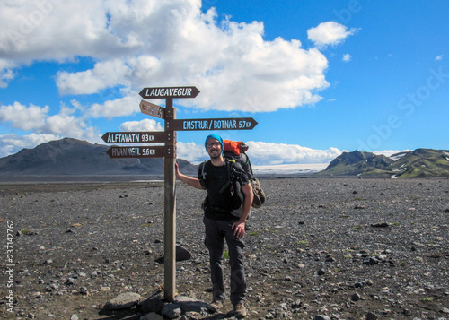 Experienced hiker with heavy backpack staying next to navigation signpost and enjoying beautiful scenery of volcanic mountains and glaciers, Iceland photo