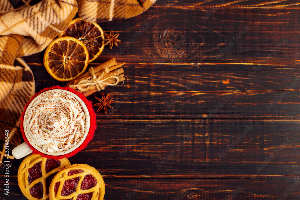 A cup of hot drink with whipped cream and powder, in a knitted cover and homemade cookies, Cezve and spices, lie on a wooden table near the checkered rug. Copy space. A cup of hot drink with whipped cream and powder, in a knitted cover and homemade cookies, Cezve and spices, lie on a wooden table near the checkered rug. Copy space.